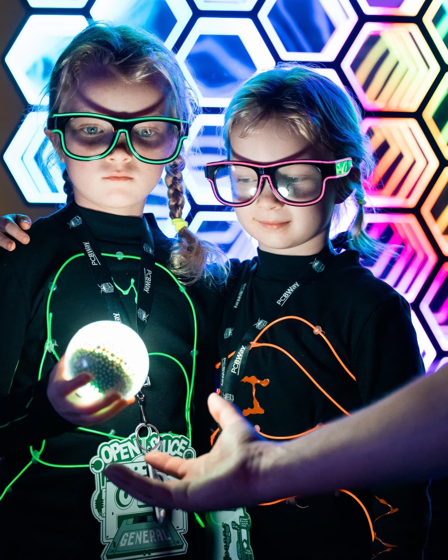 The two real sisters at Open Sauce in their glowing wire suits — one traced in green light, one in ember-orange — studying the glowing orb they built