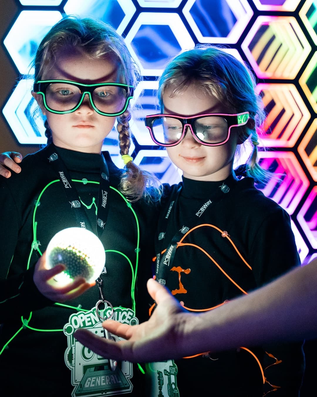 Two real sisters in glowing electroluminescent wire suits holding a bright orb of light at Open Sauce